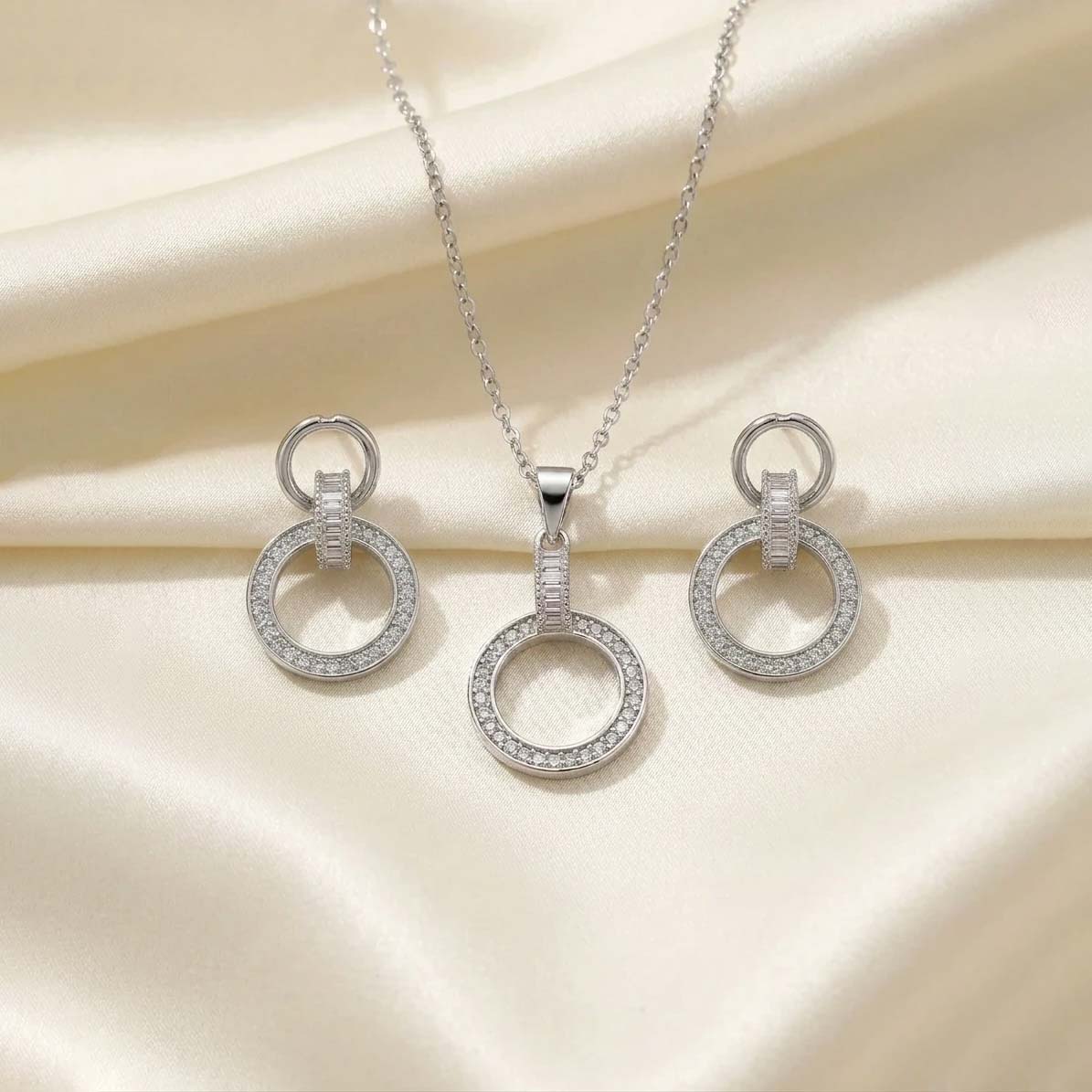Double-Circle-Silver-color-jewel-set نیمست جواهرات نقرهای رنگ شامل گردنبند ظریف و یک جفت گوشواره با طرح دو حلقه متصل به هم نگیندار که روی پارچه حریر روشن قرار گرفته است