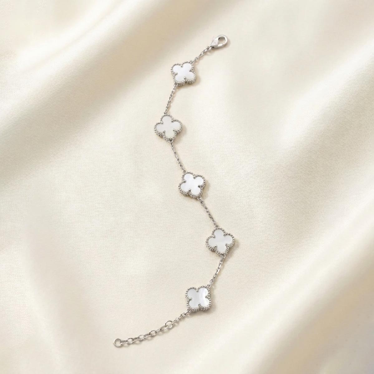 Van-Cleef-white-Clover-Bracelet-silver-color دستبند زنانه نقرهای رنگ ظریف دارای پنج آویز طرح گل شبدر چهاربرگ (ون کلیف) با داخل سفید رنگ (صدفی) و دورکاری مهرهای ظریف، قرار گرفته روی پارچه ساتن روشن.