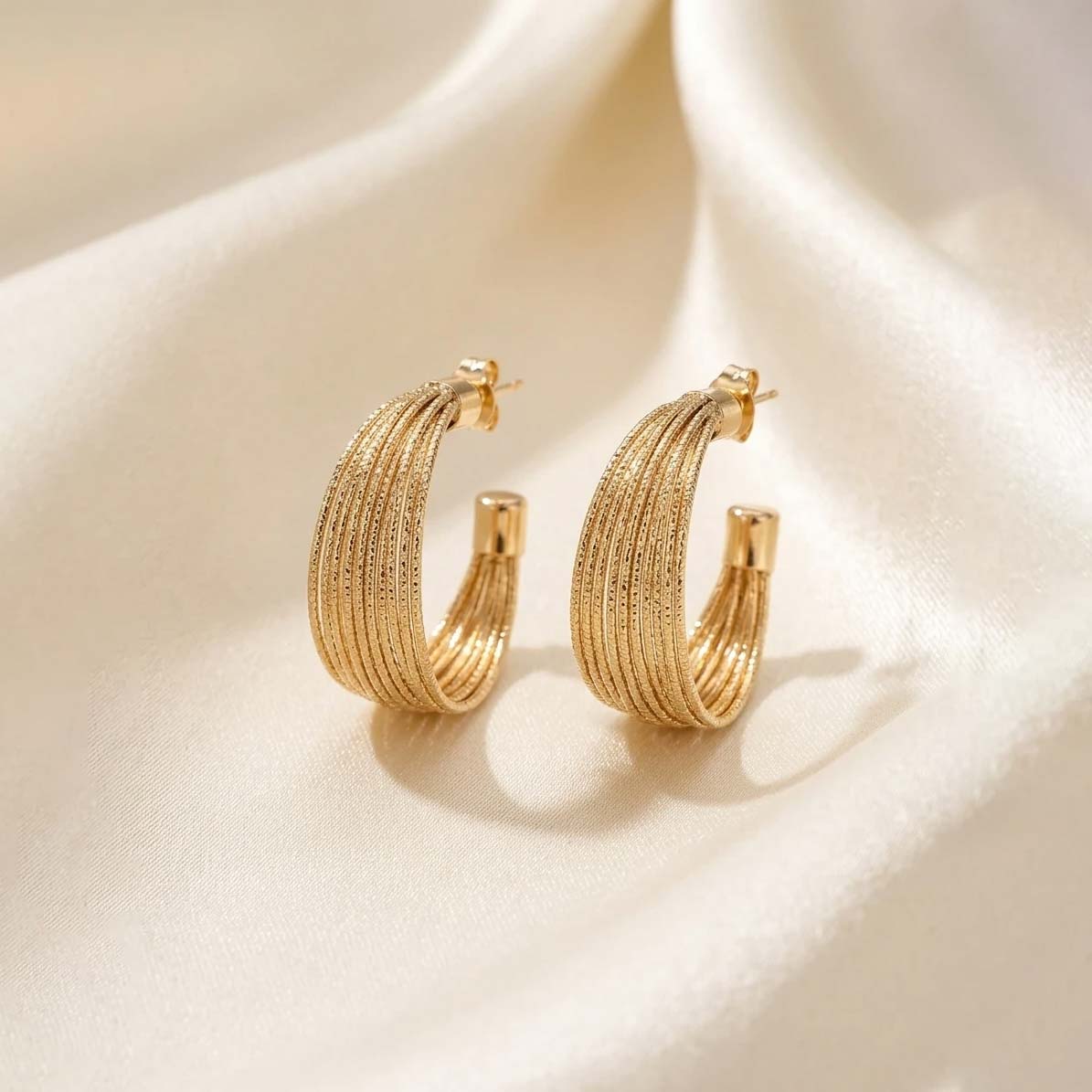 Wide-Ribbed-Gold-Hoop-Earrings یک جفت گوشواره حلقهای و تراش خورده(بافتدار) طلایی رنگ که روی پارچه حریر روشن قرار گرفته است