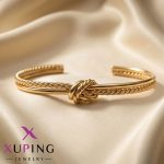 دستبند النگویی طلایی رنگ برند ژوپینگ (Xuping) با طرح گره در وسط و بازوی ترکیبی (یک سمت ساده و یک سمت مارپیچ طنابی)، قرار گرفته روی پارچه ساتن کرم.