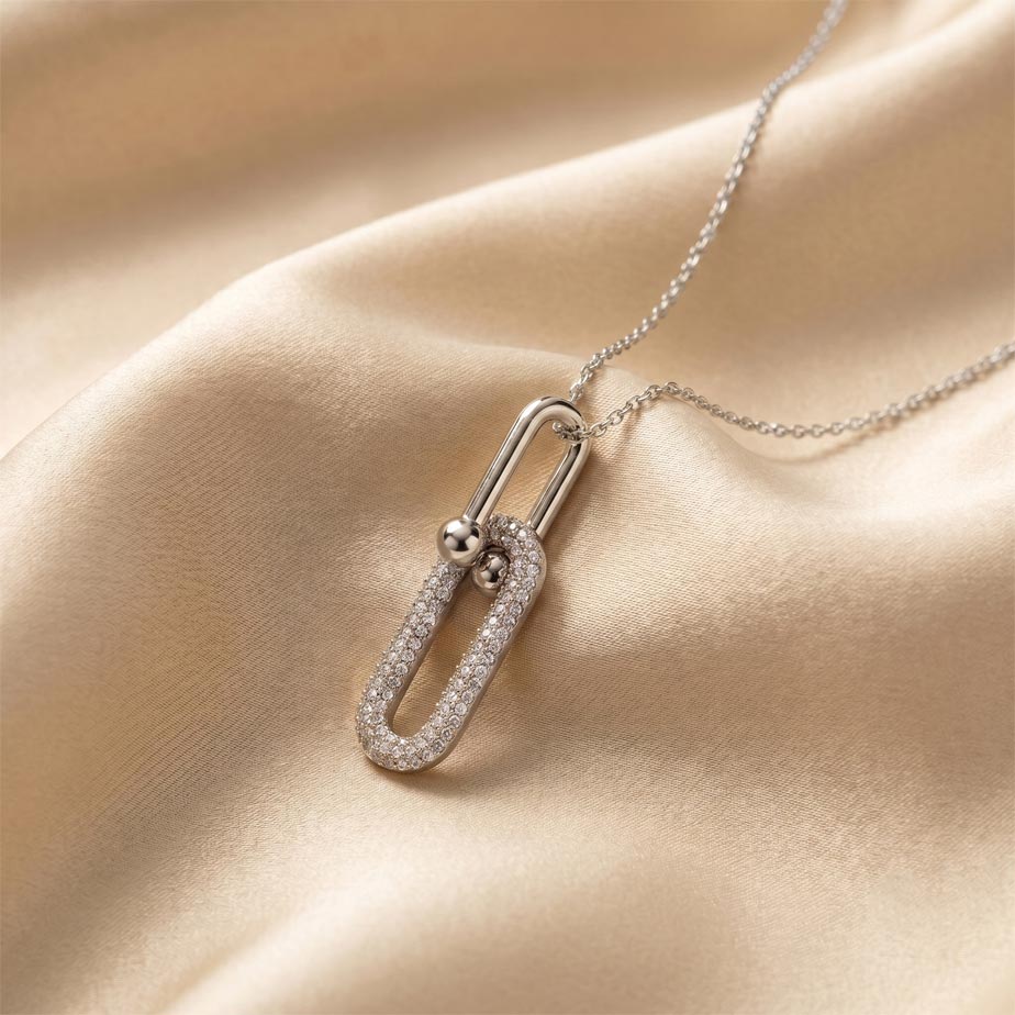 womens-silver-tiffany-style-link-necklace گردنبند نقرهای رنگ با پلاک متشکل از دو حلقه بیضی متصل به هم که حلقه پایینی با نگینهای ریز تزئین شده و روی پارچه ساتن کرم رنگ قرار دارد، نمایشی از زیبایی گردنبند نقرهای طرح تیفانی.