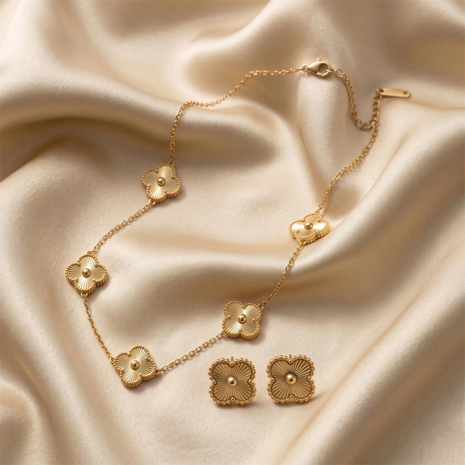 gold-van-cleef-sunburst-half-set زیورآلات طلایی رنگ شامل یک گردنبند با پنج پلاک شبدر و یک جفت گوشواره میخی. سطح پلاکهای این نیم ست طرح ون کلیف طلایی دارای خطوط شیاردار و بافت خورشیدی است که با ظرافت روی یک پارچه ساتن کرم رنگ قرار گرفتهاند.