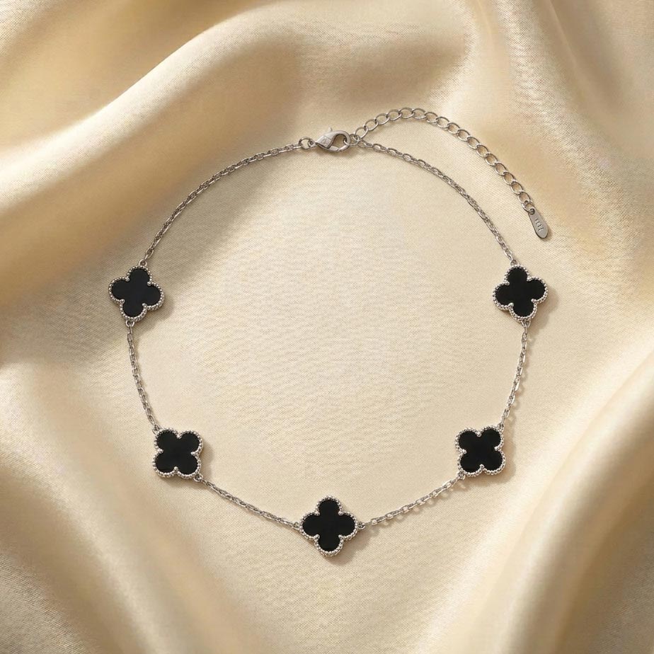 silver-necklace-van-cleef-five-motif-black-onyx گردنبند طرح ون کلیف پنج گل نقرهای قرار گرفته روی پارچه ساتن روشن با پنج پلاک به شکل شبدر چهار برگ که داخل آنها با رنگ مشکی پر شده و حاشیه دالبری ظریف دارند.